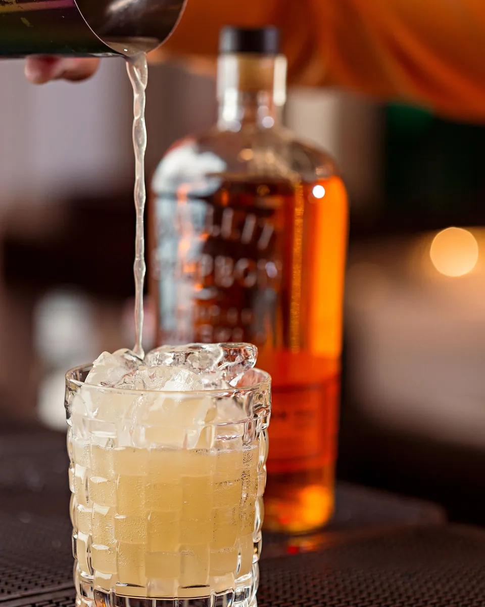 Cocktail versato al banco, bottiglia di distillato sullo sfondo